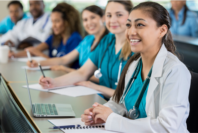 A woman wearing scrubs, a lab coat, and stethoscope smiles in a medical school classroom.
