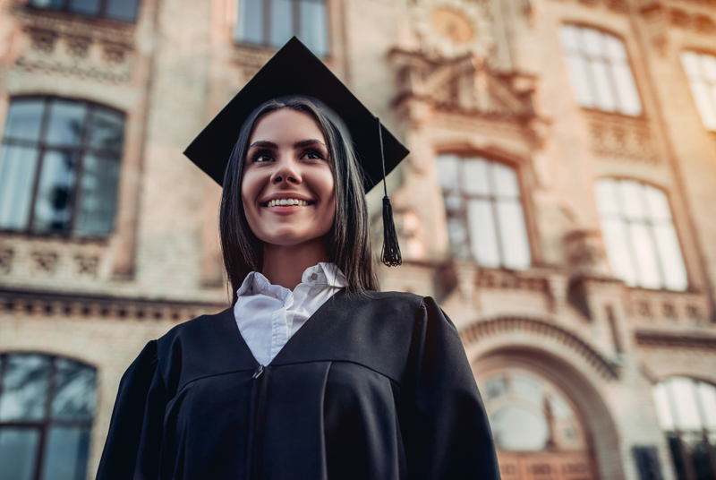 Smiling woman wearing cap and gown standing in front of university building.