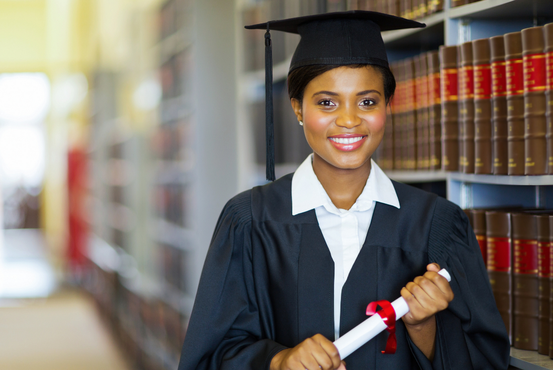 Female graduate in a cap and gown holding a diploma stands smiling near a shelf of law books.